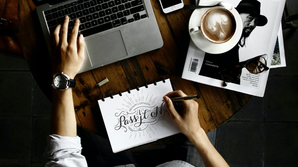 Homem visto de cima, sentado em uma mesa, experimentando um dos hobbies para desenvolver - caligrafia e lettering - escrevendo em uma folha com uma mão e mexendo em um notebook com a outra.