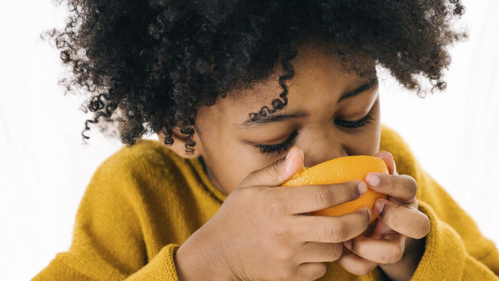 Criança de cabelo cacheado come uma laranja com as mãos, representando a boa alimentação, tema citado na palestra sobre saúde mental de Mari Luz.