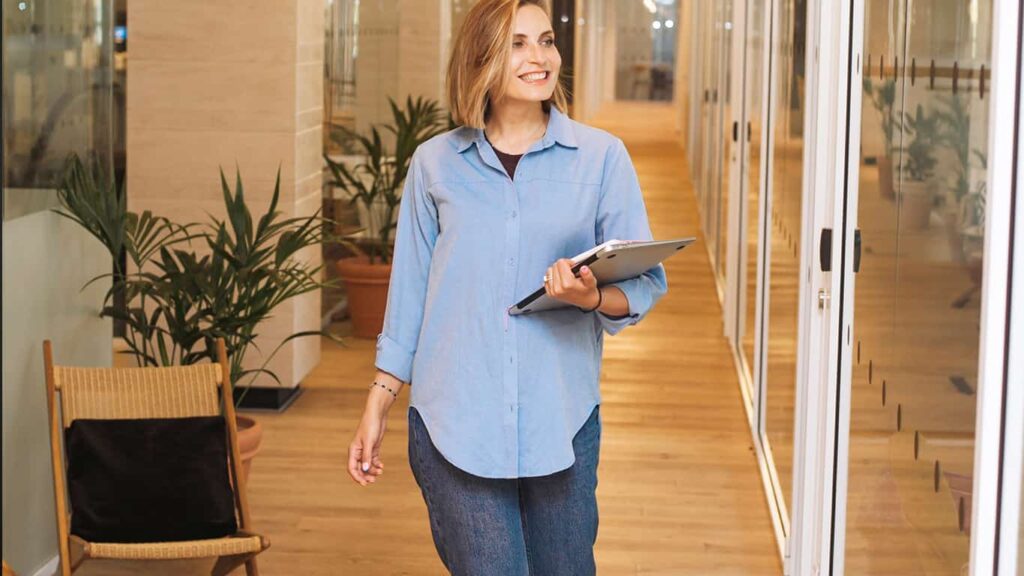 Uma mulher loira anda por um escritório sorrindo, segurando um notebook nos braços. Ela representa a modernidade e profissionalismo das palestras do MBX. 