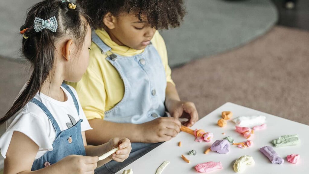 Duas crianças manuseiam massinhas coloridas, ajoelhadas em frente a uma mesa branca, em uma experiência de edutretenimento. 