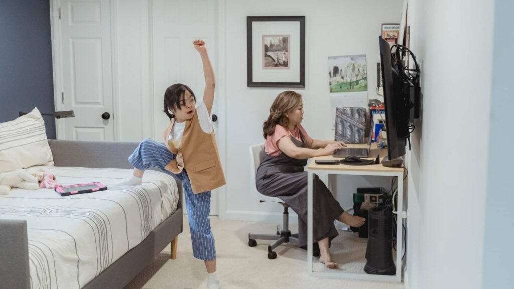 Mulher trabalha um um computador dentro de um quarto de sua casa, enquanto uma criança brinca ao lado dela com um pé em cima da cama.
