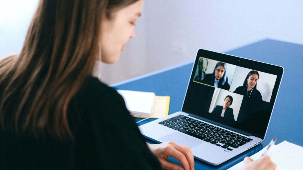Mulher sentada a uma mesa, com cara de feliz, olhando para um notebook durante uma videoconferência.