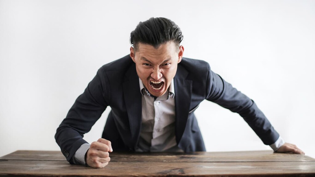 Homem de terno batendo em uma mesa com os punhos serrados, expressando uma linguagem corporal agressiva e de raiva e estresse.