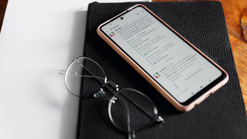 Celular e óculos na mesa. Na tela do celular, uma página com escritos longos, representando o uso saudável e consciente do celular.