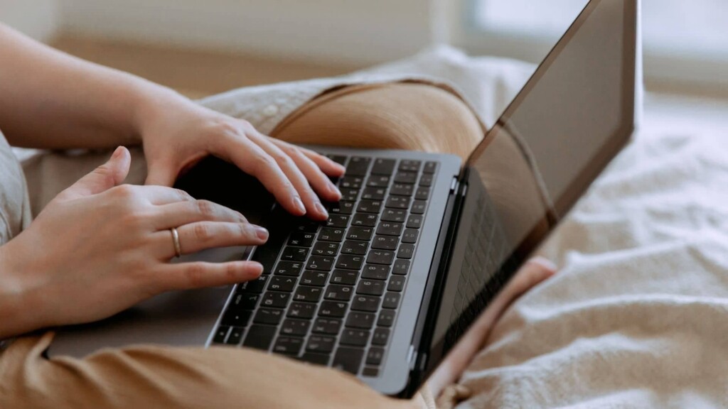Pessoa sentada em uma cama com as pernas cruzadas e um notebook em cima delas, enquanto digita.