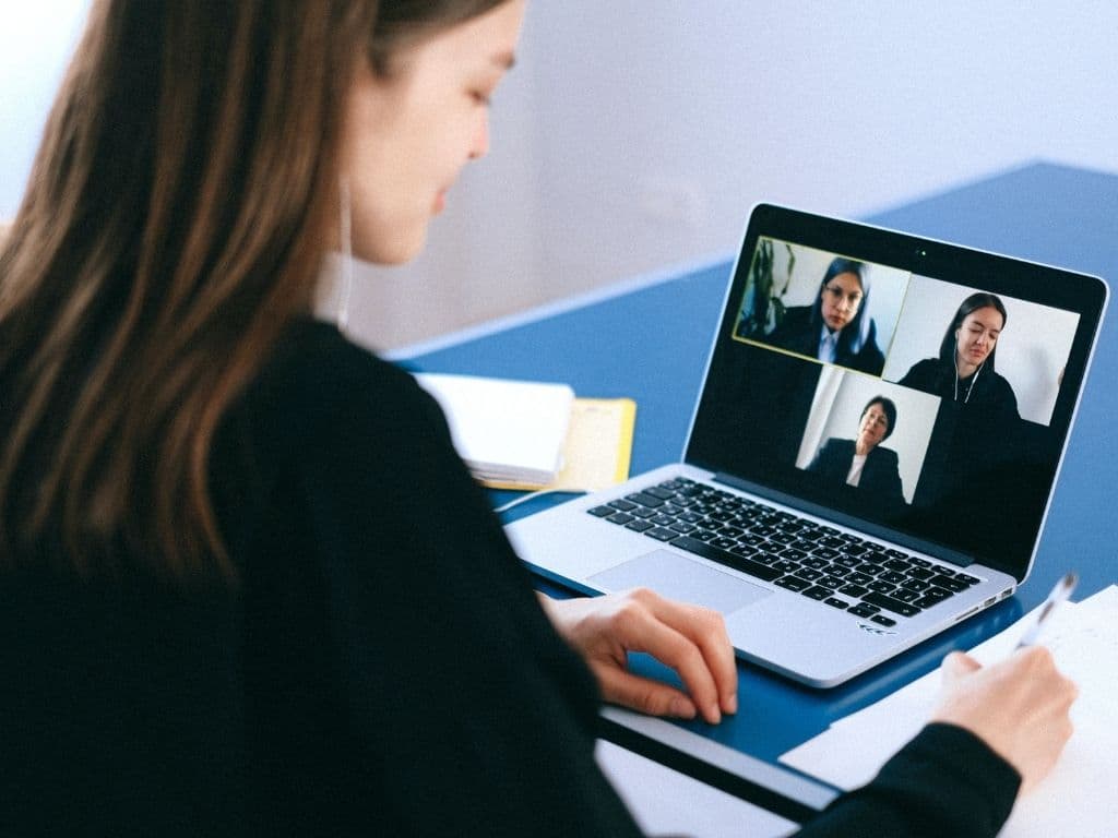 Mulher sentada a uma mesa, com um notebook ligado no qual ela participa de uma reunião online com outras três pessoas.