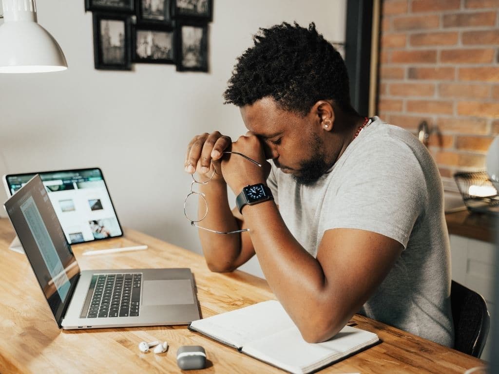 Homem sentado a uma mesa com um notebook, um tablet e um caderno abertos ao seu lado. Ele segura um par de óculos e aperta o rosto, demonstrando estar extremamente preocupado e cansado.