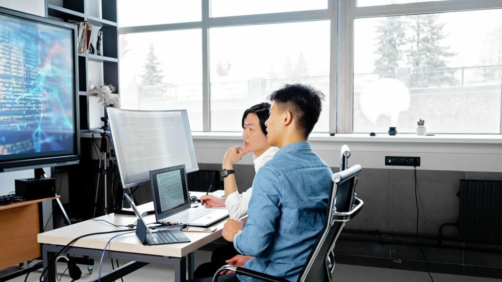Dois homens que trabalham com Digital Business sentam a uma mesa, utilizando com um computador e um tablet, enquanto olham para uma televisão com informações de negócios e conversam.