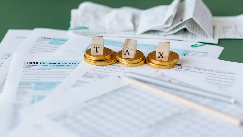Papéis de gestão tributária em cima de uma mesa, com algumas moedas em cima deles e pequenos blocos de madeira soletrando a palavra "Tax" (que significa imposto, em inglês).