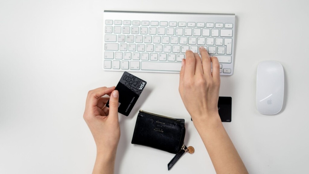 Mão de uma pessoa segurando um cartão de crédito preto, enquanto a outra mão digita em um teclado de computador.
