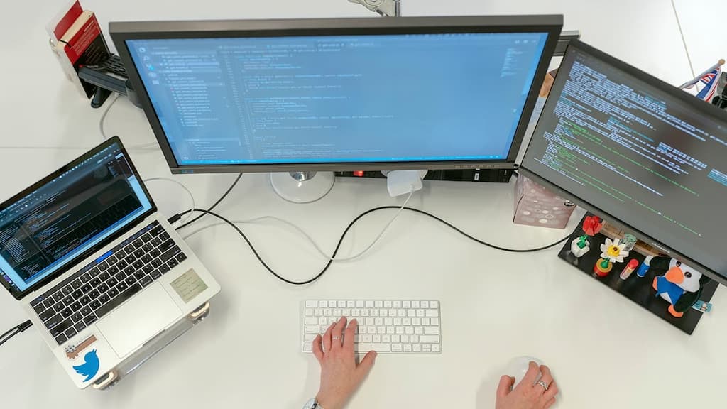 Três computadores vistos de cima, trabalhando com Engenharia de Dados. Uma mão no teclado e uma mão no mouse. 