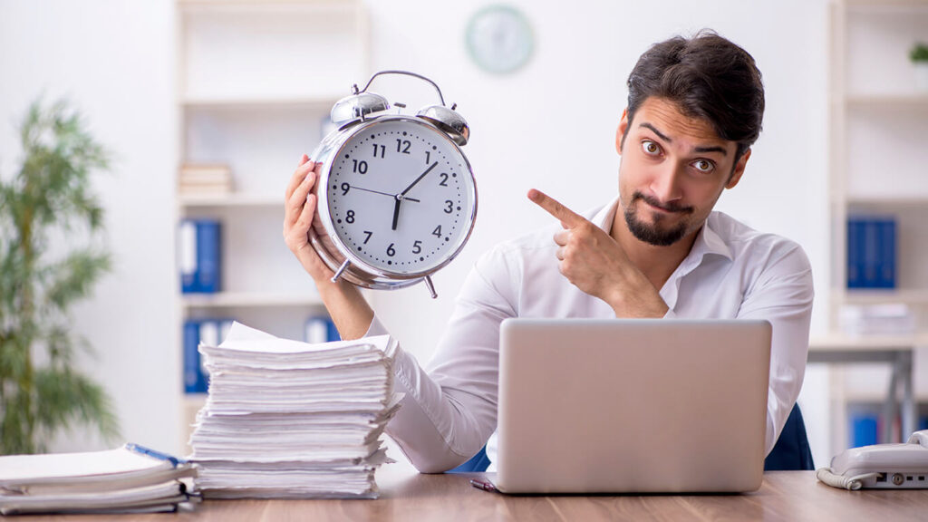 Homem sentado em frente ao computador, apontando para um relógio, representação da gestão do tempo