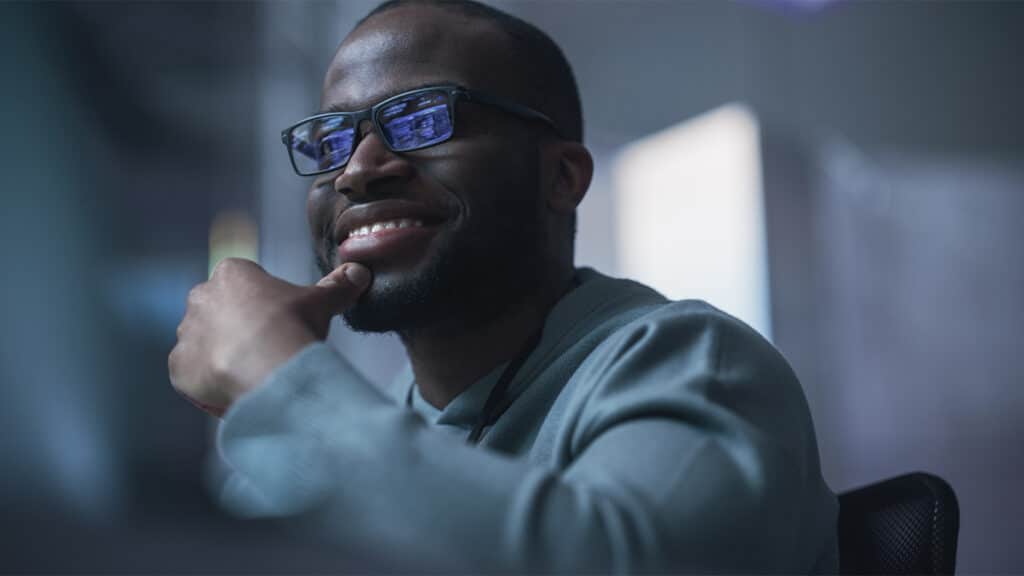 Homem negro, careca, usando moleton claro, de óculos, com a mão no queixo, olhando para uma tela de computador em enquadramento frontal, com expressão de riso e felicidade.