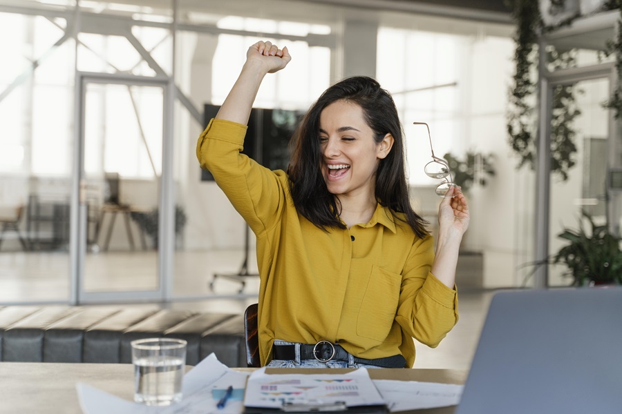 Em um escritório, após entender como se destacar no trabalho, uma jovem mulher branca comemora uma conquista profissional; ela está sentada à mesa, em frente a computador e relatórios.