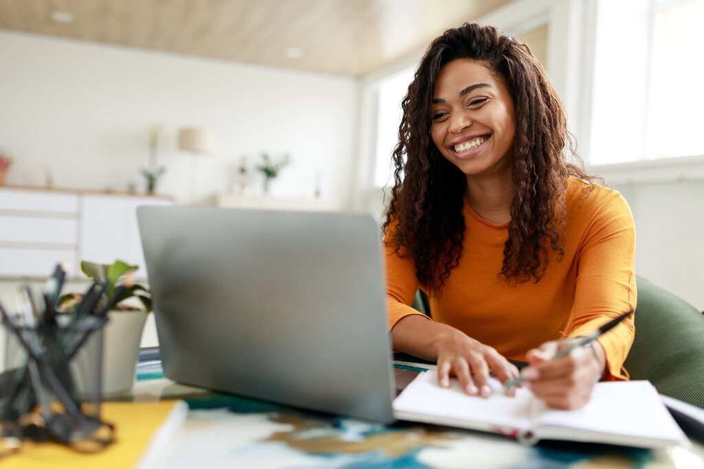 Mulher sorridente, utilizando caderno e notebook para participar de curso de especialização.