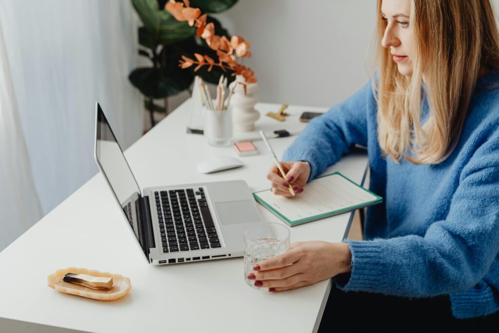 Pessoa trabalhando em seu escritório doméstico, escrevendo em uma agenda enquanto utiliza um laptop, ilustrando o foco e a organização necessários no desenvolvimento de habilidades de liderança.