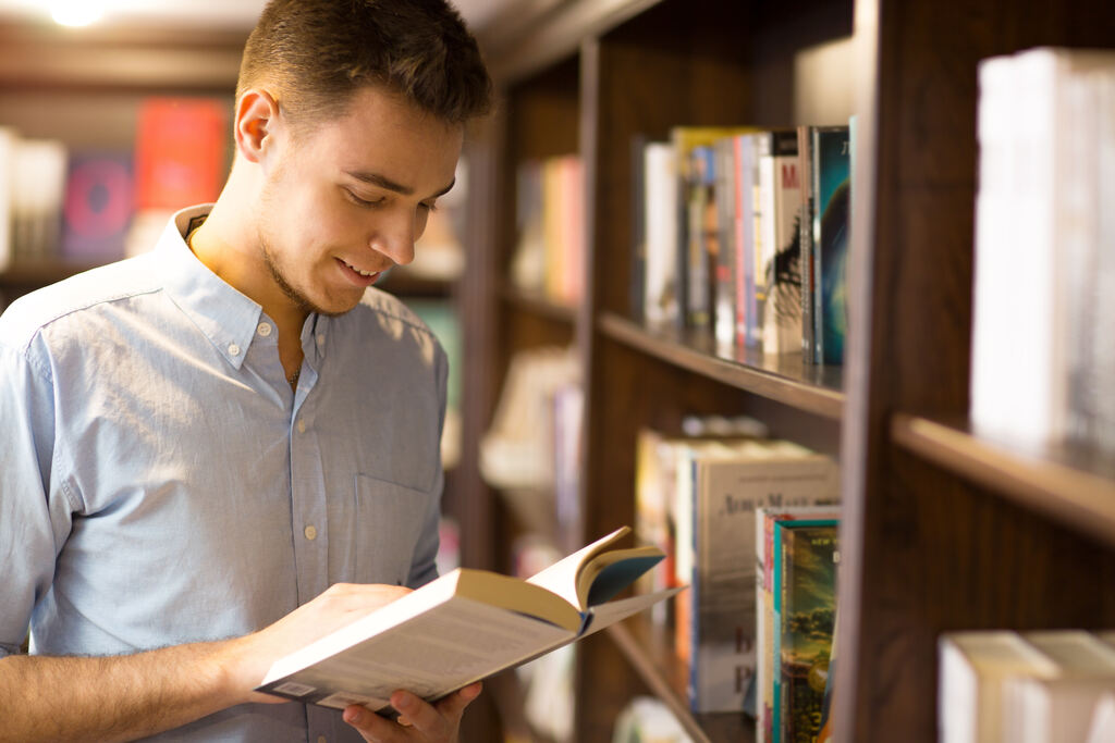 Jovem homem em biblioteca, utilizando livro para pesquisa de disciplinas de cursos de especialização.