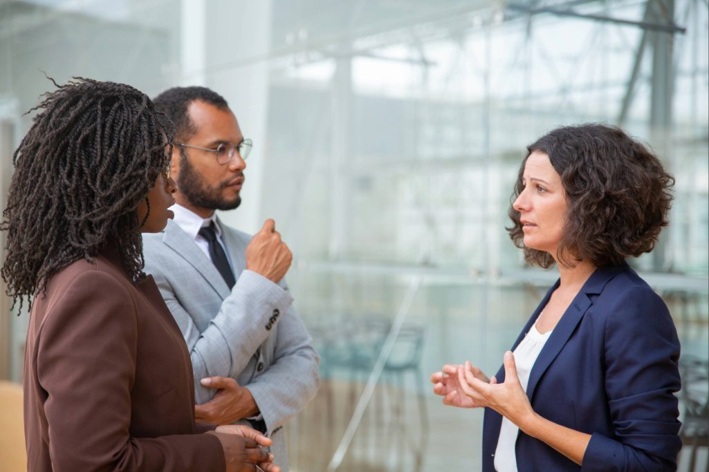 Três colegas de trabalho, de diferentes etnias, participam de uma conversa profissional, demonstrando comunicação assertiva no ambiente corporativo.