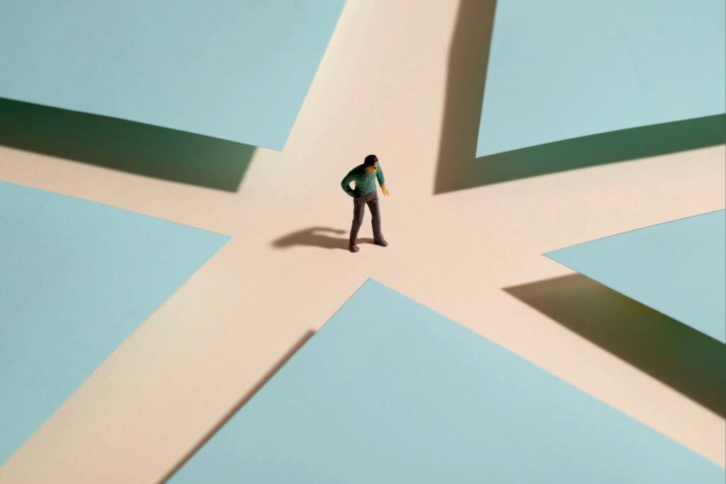 Imagem ilustrando uma pessoa em um ponto de decisão, representada por uma miniatura no centro de várias folhas de papel azul dispostas em forma de cruz.