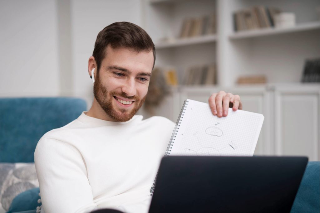 Homem sorrindo e olhando para um notebook que está em seu colo, sentado em um sofá.