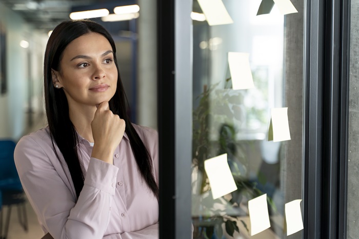 Uma profissional está olhando fixamente para um mural de vidro com post-its;ela parece avaliar opções, como se fosse fazer uma mudança de carreira.